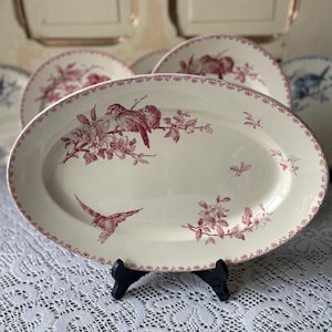 Peut inclure: Un plat de service ovale avec une base crème et un motif floral et d'oiseaux bordeaux. Le bord est orné d'un motif bordeaux délicat. Le plat est soutenu par un support noir.