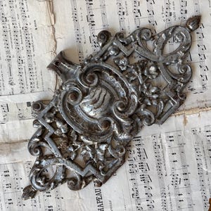 Puede incluir: Adorno arquitectónico antiguo de metal plateado. El elemento decorativo presenta diseños florales y de volutas. Descansa sobre un fondo de partituras antiguas.