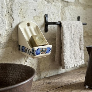 Può includere: Un portasapone in ceramica color crema con decorazioni floreali blu e una saponetta. Un portasciugamani in metallo nero sostiene un asciugamano beige. Gli elementi sono montati su un muro di pietra strutturato, ambiente rustico.