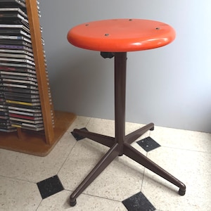 Peut inclure: Un tabouret doté d'une assise circulaire orange vif et d'une colonne centrale marron reposant sur une base à quatre pieds. L'assise est fixée à la colonne par des vis métalliques visibles, et la base comporte de petits pieds noirs.