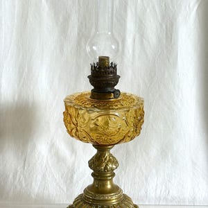 Peut inclure: Lampe à huile ancienne avec une cheminée en verre transparent et un réservoir en verre ambré décoratif. La lampe présente une base en laiton ornée et des motifs en relief détaillés, mettant en valeur une esthétique vintage.