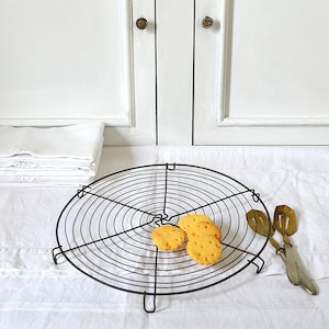 May include: A black wire cooling rack holds several golden-brown cookies. A pair of vintage serving tongs rests beside the rack on a white tablecloth. A stack of folded white linens and a white cabinet are in the background.