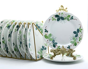 May include: A set of white ceramic plates featuring a botanical strawberry plant pattern with green leaves, small white flowers, and red berries. The plates are organized in a gold-toned wire rack, with one plate displayed on a decorative gold stand.