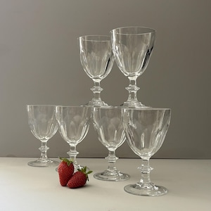 Peut inclure: Un ensemble de six verres à pied en verre transparent avec des détails cannelés verticaux sur les bols et des pieds à facettes, disposés en formation étagée. Deux fraises rouge vif avec des feuilles vertes sont placées devant les verres.