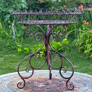 May include: A brown metal plant stand featuring a circular basket top with a lattice wire design and a decorative rim. The base consists of three curved legs with intricate scrollwork details, providing a stable structure for the elevated tray.