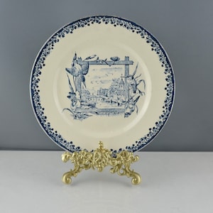 Op de afbeelding: Een vintage blauw-witte keramische plaat met een decoratief ontwerp van een klein stadje dat wordt omlijst door een raam. De plaat staat op een gouden metalen standaard.