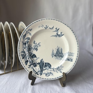 Peut inclure: Assiette en céramique crème avec un motif floral et d'oiseaux bleu. L'assiette présente une bordure de petits points bleus et une illustration détaillée d'oiseaux, de fleurs et de voiliers. Un ensemble d'assiettes est visible en arrière-plan.