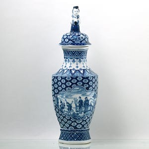 May include: A blue and white porcelain vase with a lid featuring a small figurine of a person. The vase has a decorative design with a scene of people in a landscape.