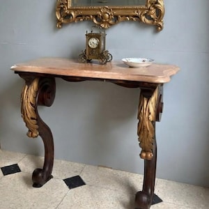 Peut inclure: Une console avec un plateau en marbre rose clair et des pieds en bois foncé ornés de détails sculptés en forme de feuilles dorées. La table possède deux pieds incurvés se terminant par des volutes. Une horloge et un bol sont posés dessus.