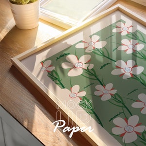 Puede incluir: Una impresión enmarcada con un fondo verde y flores blancas con centros rojos. La impresión está en un marco de madera claro y el texto "Mock Up Paper" es visible en la esquina inferior derecha.