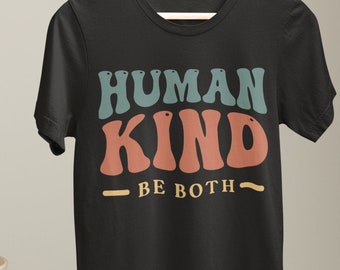 Puede incluir: Camiseta negra con el texto "HUMAN KIND BE BOTH" en letras de estilo retro. El texto es de color turquesa y coral. La camiseta está colgada de una percha de madera. Jarrones decorativos y un pedestal en el fondo.