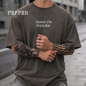 Könnte beinhalten: Ein anthrazitfarbenes Kurzarm-T-Shirt mit Rundhalsausschnitt. Auf der Brust befindet sich der weiße Schriftzug 'Armor On Everyday' und das Wort 'PEPPER' in der oberen linken Ecke. Der Stoff hat eine verwaschene Vintage-Optik und eine lockere Passform.