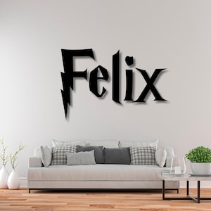 Könnte beinhalten: Schwarze Wandkunst mit dem Namen "Felix" in einer stilisierten Schriftart mit einem Blitz. Der Name wird an einer weißen Wand über einem weißen Sofa mit Kissen, einem Couchtisch und einer Pflanze angezeigt.