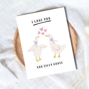 May include: A white greeting card featuring the text "I LOVE YOU" above an illustration of two geese with blue bows. Pink hearts float between the geese, and the text "YOU SILLY GOOSE" is below. The card rests on a wooden plate.