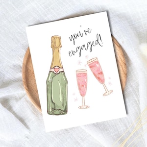 May include: A greetings card featuring a champagne bottle and two glasses of pink champagne. The words "you're engaged!" are written in black script. The card is on a wooden tray.