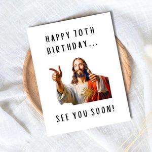 以下が含まれることがあります： 「HAPPY 70TH BIRTHDAY... SEE YOU SOON!」のテキストが入った白いグリーティングカード。親指を立てて指を指しているイエスの画像が特徴です。カードは木製のプレートと白い布の上にあります。