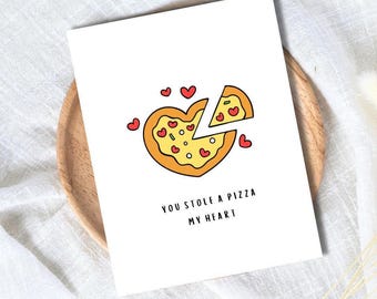 Op de afbeelding: Een witte wenskaart met een hartvormig pizzadesign. De pizza heeft een gele korst, rode hartvormige toppings en de tekst "YOU STOLE A PIZZA MY HEART". De kaart ligt op een houten bord.