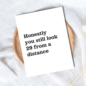Op de afbeelding: Een witte kaart met de tekst "Honestly you still look 29 from a distance" in het zwart. De kaart ligt op een houten bord, tegen een witte stoffen achtergrond.
