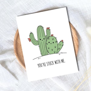 May include: A white greeting card featuring a cartoon illustration of two green cacti with smiling faces and small pink flowers. The text below the cacti reads, "YOU'RE STUCK WITH ME." The card has a clean, simple design with black outlines.