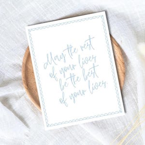May include: A white card with a light blue scalloped border and text that reads, "May the rest of your lives be the best of your lives." The card is on a wooden plate, set against a white cloth.