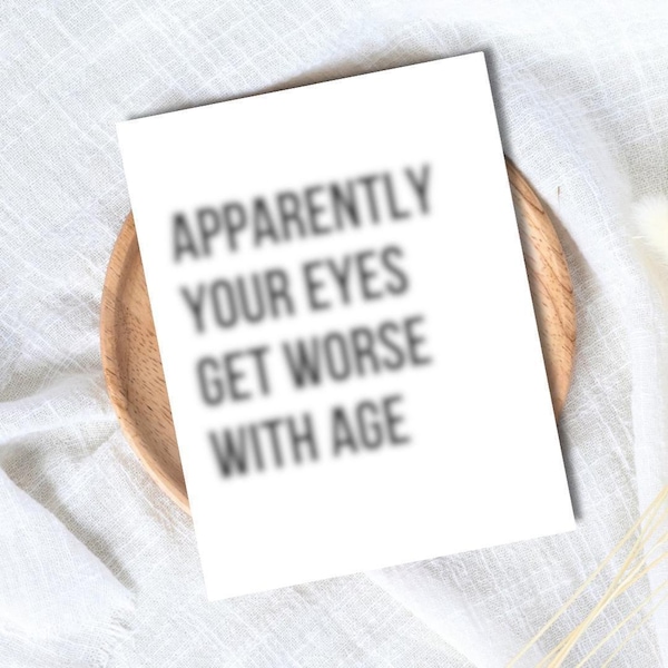 以下が含まれることがあります： 「Apparently Your Eyes Get Worse With Age」の黒い文字が書かれた白いグリーティングカード。木製のプレートの上に置かれ、白い布の背景に配置されています。テキストはわずかにぼやけています。