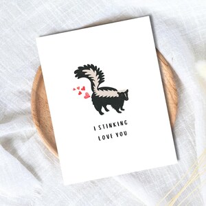 May include: A white greeting card featuring a cartoon skunk with a black and white body, a fluffy tail, and red heart accents. The text "I STINKING LOVE YOU" is printed below the skunk in black font. The card is on a wooden plate.