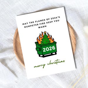May include: A white greeting card featuring a green dumpster with the text 2026, containing a burning Christmas tree with orange flames. The text reads: MAY THE FLAMES OF 2026'S DUMPSTER FIRE KEEP YOU WARM and merry christmas in green script.