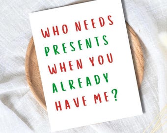 May include: A white greeting card with the text "WHO NEEDS PRESENTS WHEN YOU ALREADY HAVE ME?" in red and green lettering. The card is on a wooden plate, set against a white, textured background.