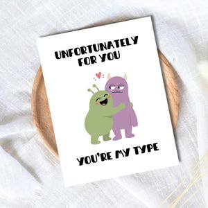 May include: A white greeting card featuring a cartoon illustration of a green alien hugging a purple monster. Above the characters, the text reads "UNFORTUNATELY FOR YOU" and below them, the text reads "YOU'RE MY TYPE" in bold black lettering.