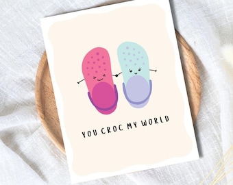 Könnte beinhalten: Eine Grußkarte mit hellbeigem Hintergrund zeigt zwei Cartoon-Clogs, einen rosa und einen blauen, die sich an den Händen halten. Der Text "YOU CROC MY WORLD" ist darunter in Schwarz gedruckt. Die Karte liegt auf einem Holztablett.