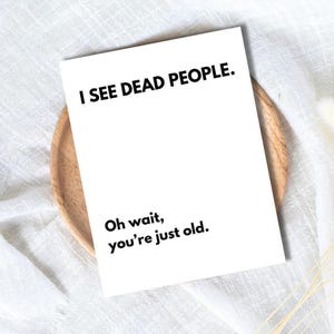 May include: A white greeting card with the text "I SEE DEAD PEOPLE." and the text "Oh wait, you're just old." The card is on a wooden plate, set against a white fabric background.