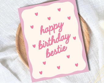 Puede incluir: Tarjeta de felicitación rosa y crema con las palabras "happy birthday bestie" en escritura rosa. Pequeños corazones rosas están esparcidos alrededor del texto. La tarjeta está sobre un plato de madera.