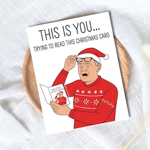 Peut inclure: Carte de Noël humoristique avec le texte « THIS IS YOU... TRYING TO READ THIS CHRISTMAS CARD ». L'illustration montre un homme portant un bonnet de Père Noël et un pull rouge, peinant à lire une petite carte.