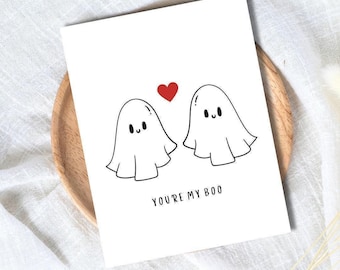 May include: A white greeting card with a black and red illustration. The card features two cartoon ghosts with a red heart above them and the text "YOU'RE MY BOO" below. The card is on a wooden plate.