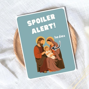 May include: A greeting card with a light blue background features a religious illustration of the Holy Family. The text "SPOILER ALERT!" is at the top, with "he dies" and an arrow pointing to the baby. The card is on a wooden plate.