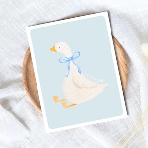May include: A watercolor illustration of a white goose with a blue ribbon around its neck, set against a light blue background. The words "LITTLE ONE" are written in a script font. The artwork is on a white rectangular card.