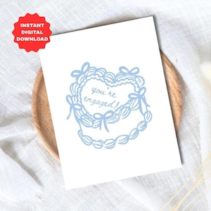 Könnte beinhalten: Eine weiße rechteckige Grußkarte mit einer hellblauen Illustration einer herzförmigen Torte mit gerüschtem Zuckerguss und Schleifen. Der Text "you're engaged!" steht in der Mitte der Torte in einer hellblauen Schreibschrift.