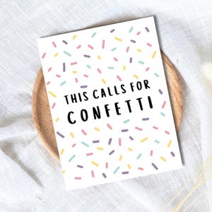 Könnte beinhalten: Eine weiße Grußkarte mit bunten Konfetti-ähnlichen Streuseln und dem Text "THIS CALLS FOR CONFETTI" in Schwarz. Die Karte liegt auf einem Holztablett vor einem weißen Stoffhintergrund.