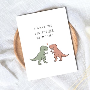 May include: A white greeting card featuring a cartoon illustration of a green Tyrannosaurus Rex and a pink Tyrannosaurus Rex holding a small flower. Above the dinosaurs, the text reads I WANT YOU FOR THE REX OF MY LIFE with a small heart.