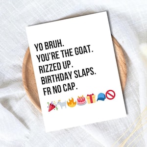 May include: A white greeting card with black text that reads "YO BRUH. YOU'RE THE GOAT. RIZZED UP. BIRTHDAY SLAPS. FR NO CAP." with several emojis. The card is on a wooden plate.