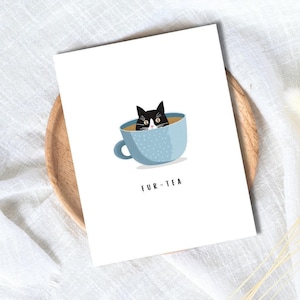 May include: A white card with a cartoon cat illustration. The cat, black and white, is in a light blue teacup with white dots. The words "FUR-TEA" are printed below the teacup. The card is on a wooden plate.