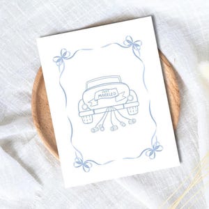 May include: A white card featuring a light blue drawing of a car with a "Just Married" banner and cans tied to the back. The card is decorated with light blue bows and ribbons. It rests on a wooden plate.