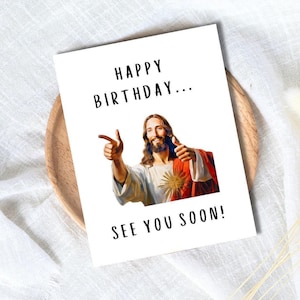 以下が含まれることがあります： 「HAPPY BIRTHDAY... SEE YOU SOON!」のテキストが入った白いバースデーカード。親指を立てて指を指しているイエスの画像が特徴です。カードは木製のプレートの上にあり、白いテクスチャの背景に配置されています。