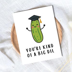 May include: A white greeting card featuring a cartoon pickle wearing a graduation cap. The pickle has a smiling face and outstretched arms. The text on the card reads, "YOU'RE KIND OF A BIG DIL."