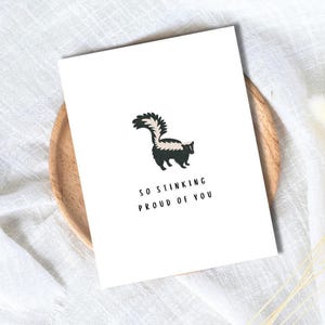 May include: A white greeting card featuring a cartoon skunk. The skunk is depicted in black and white with a bushy tail. The card has the text "SO STINKING PROUD OF YOU" printed in black beneath the skunk.
