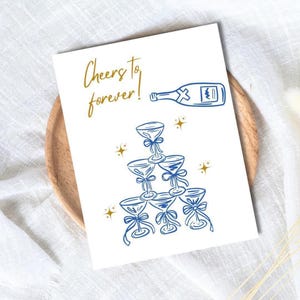 Op de afbeelding: Een witte kaart met de gouden tekst "Cheers to forever!" en een blauwe illustratie van een champagnefles en een piramide van champagneglazen. De kaart ligt op een houten dienblad.