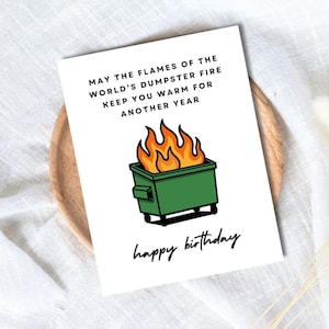 May include: A white birthday card with the text "MAY THE FLAMES OF THE WORLD'S DUMPSTER FIRE KEEP YOU WARM FOR ANOTHER YEAR" and "happy birthday". The card features a green dumpster on fire.