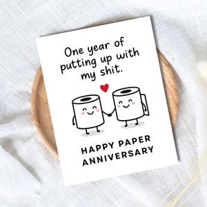 May include: A white greeting card with the text "One year of putting up with my shit." Two cartoon toilet paper rolls holding hands, with a red heart above them. The card reads "Happy Paper Anniversary."