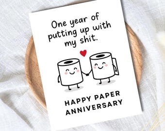 May include: A white greeting card with the text "One year of putting up with my shit." Two cartoon toilet paper rolls holding hands, with a red heart above them. The card reads "Happy Paper Anniversary."
