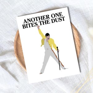 以下が含まれることがあります： 「ANOTHER ONE BITES THE DUST」の文字が書かれた白いカード。黄色いジャケットと白いジャンプスーツを着て杖を持っている人物のイラストが描かれています。このイラストは有名なミュージシャンへのオマージュです。